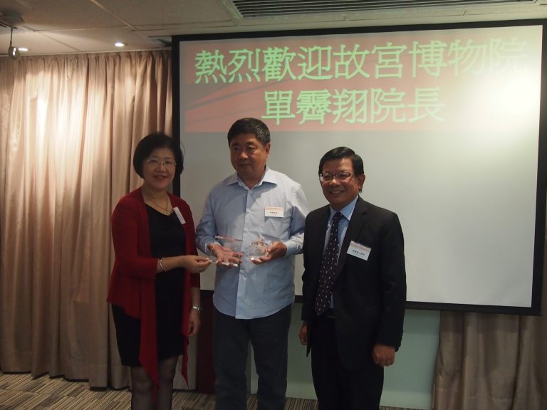 "Three people at event, one holding award, screen behind reads ""Welcome Director Chien of the Palace Museum""."